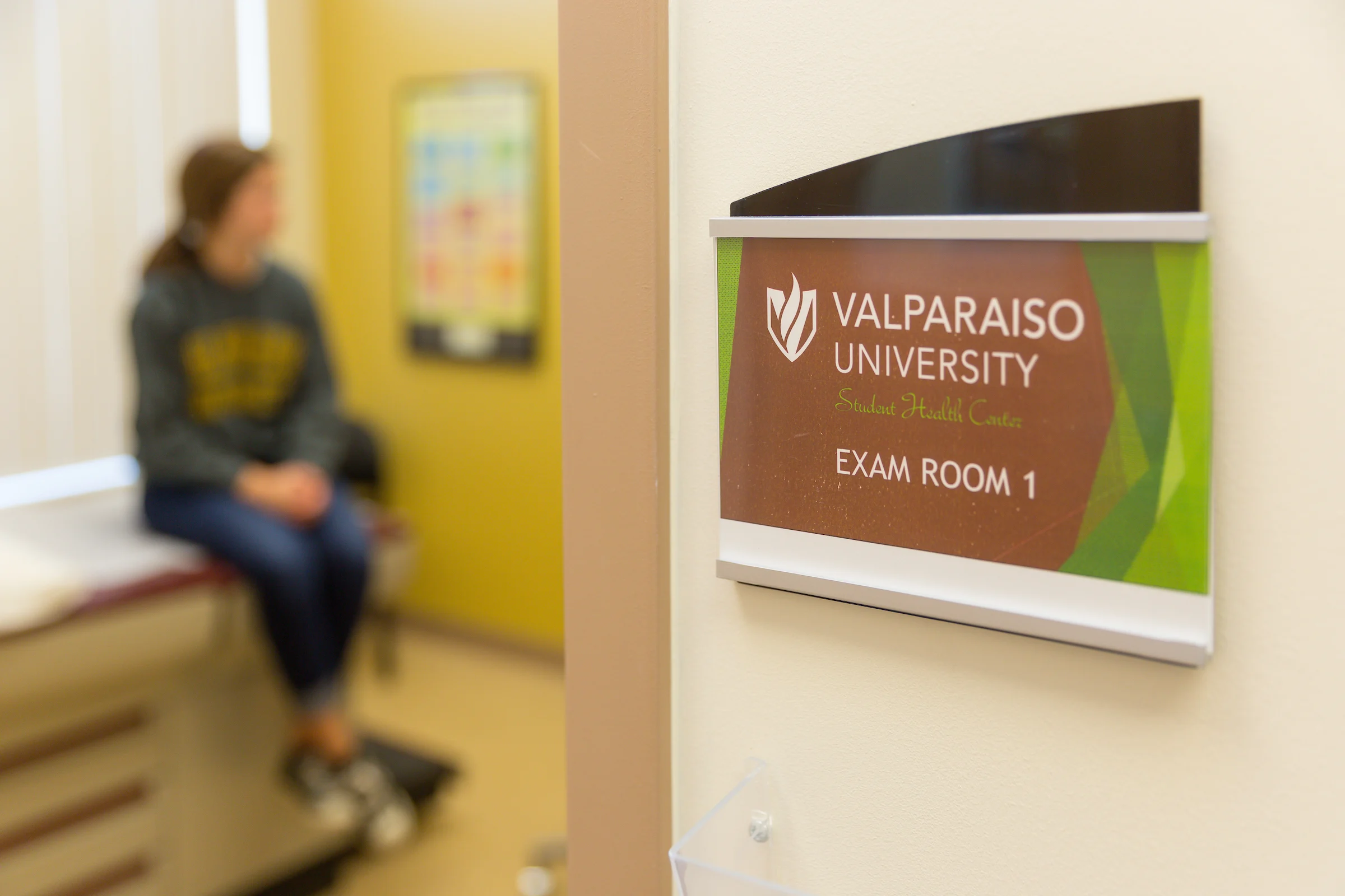 A sign for "Valparaiso University Wellness Exam Room 1" outside a medical exam room with a patient inside, promoting health and care.