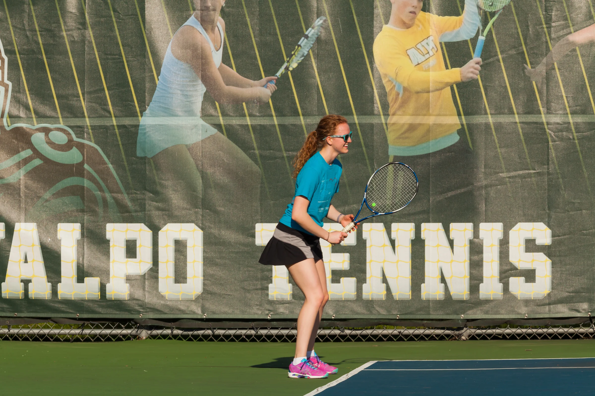 A girl in a blue shirt and black skirt prepares to receive a tennis serve on an outdoor court, embracing health and wellness through active play.