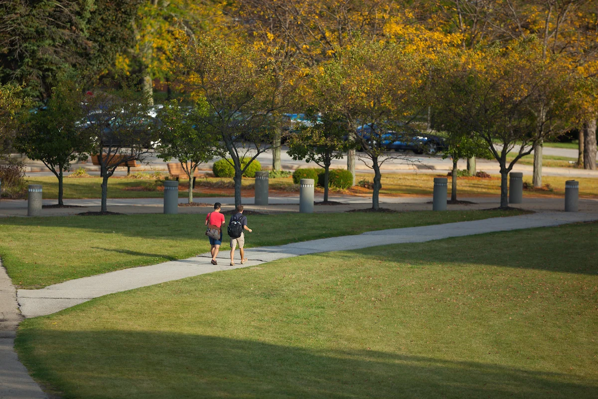 Two people walk on a campus sidewalk surrounded by grass and trees with autumn leaves—¡Porque Somos Como Tú!.