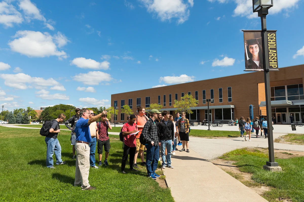 A group of students tours a college campus on a sunny day near a scholarship banner, learning about the various services available to support their academic journey.