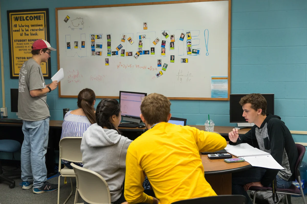 Four students work at computers while one writes on a whiteboard covered in notes and colorful cards during a peer tutoring session.
