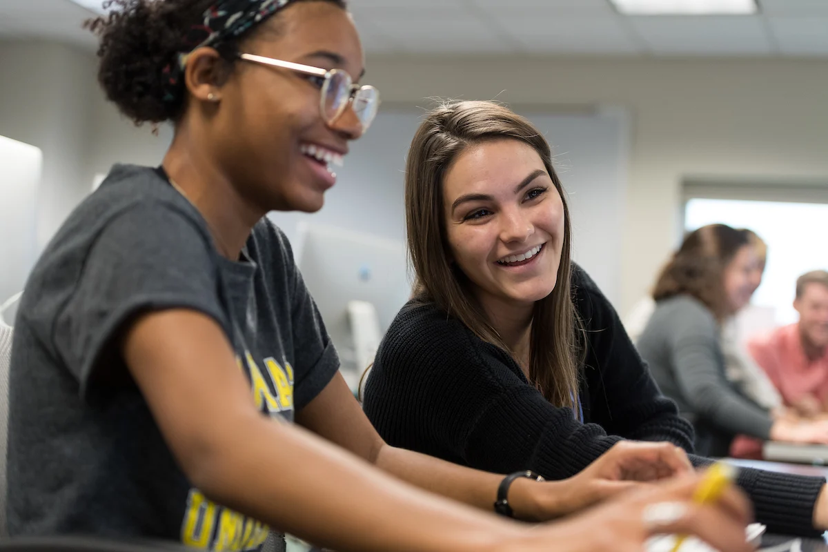Two young women, both admitted students, smiling and working together at computers in a classroom setting.