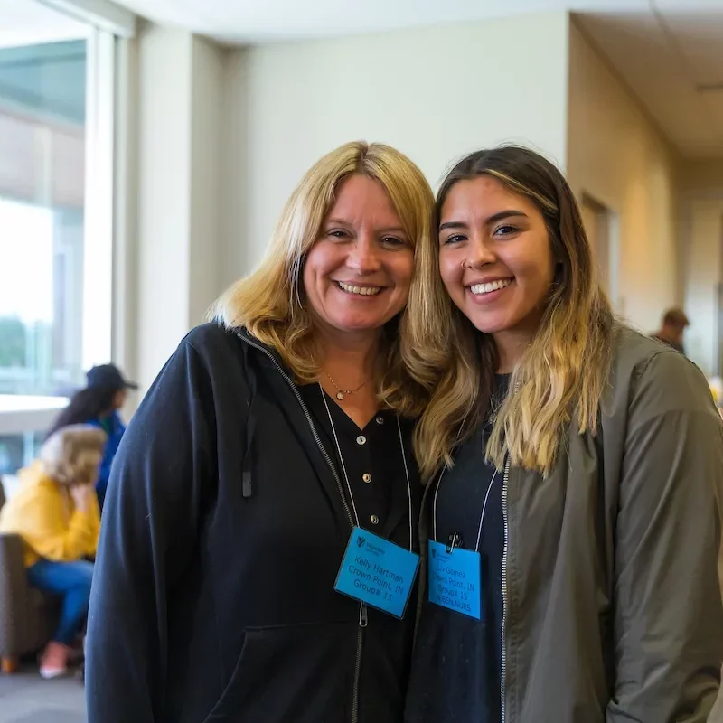 Two women smiling together indoors, both wearing jackets and blue name tags around their necks, demonstrating the spirit of HSI Principles in their friendly interaction.
