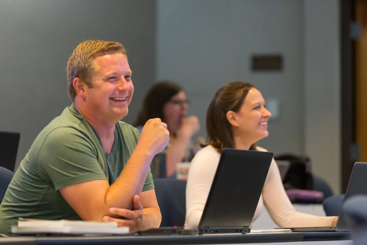 Two people sit at desks with laptops, smiling and listening attentively in a classroom setting, engaged in an EdAssist learning session.