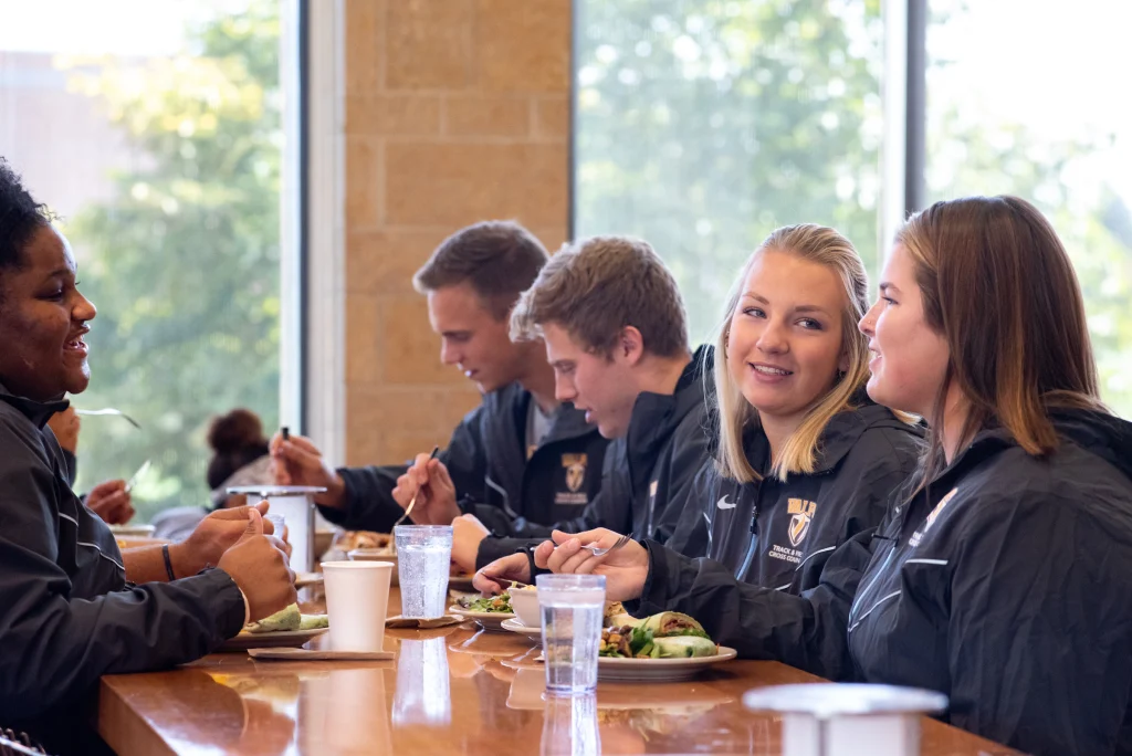 Five students in matching jackets enjoy dining and talking together at a table by a large window in a bright cafeteria.