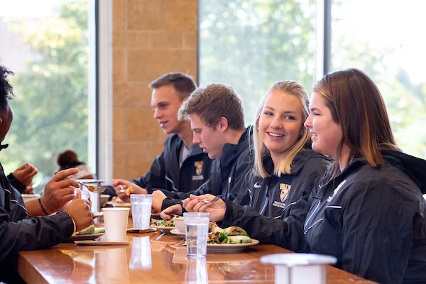Four young adults in matching jackets sit at a table, enjoying a healthy meal and lively conversation in a bright cafeteria, reflecting their commitment to health & wellness.