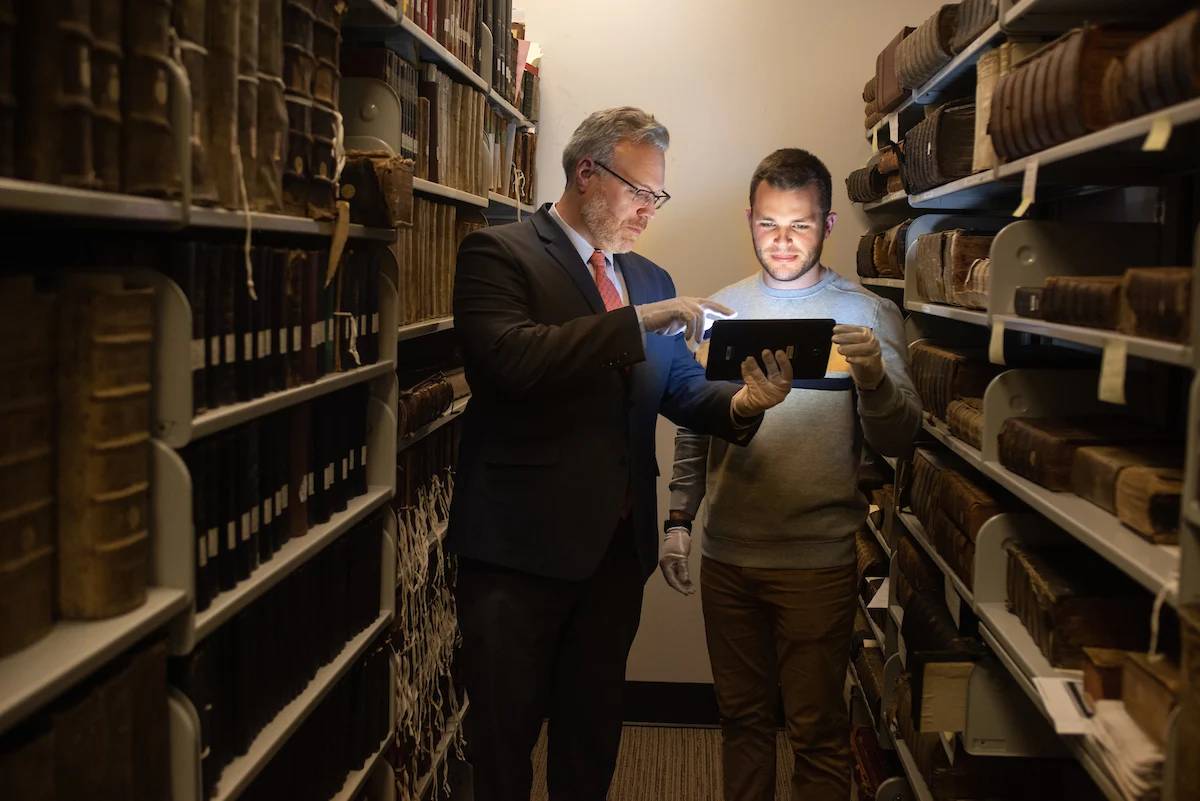 Two men stand in a library aisle, looking at a tablet surrounded by shelves of old books, discussing the new online application submission forms.