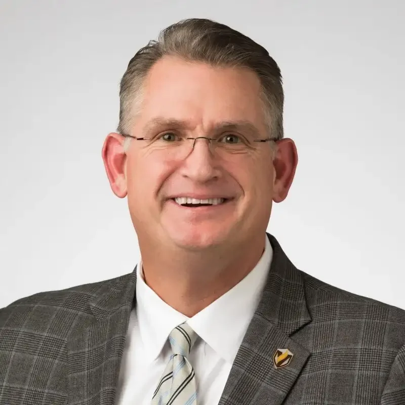 Smiling man in glasses and plaid suit jacket with striped tie, exuding confidence in communications, against a plain light background.