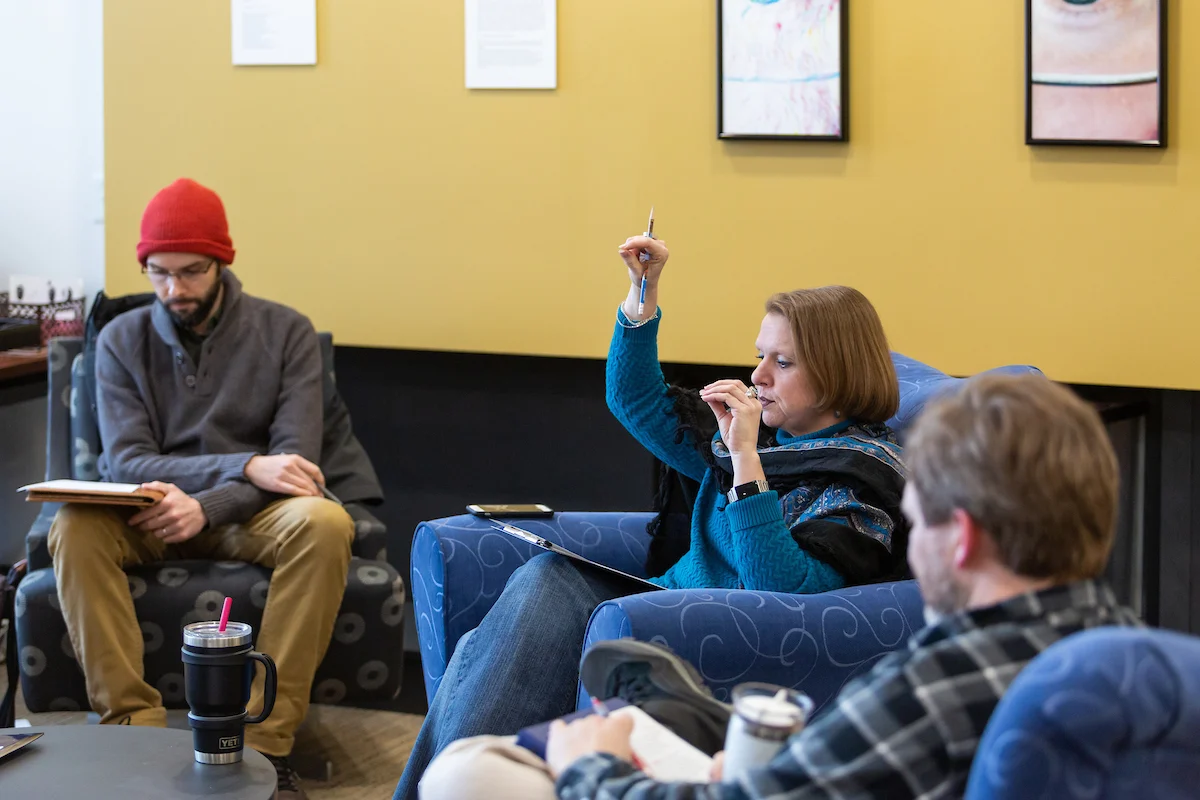 Three people sit and talk in a casual room; one woman raises her hand while the others listen, discussing improvements to their Online Application Submission Forms.
