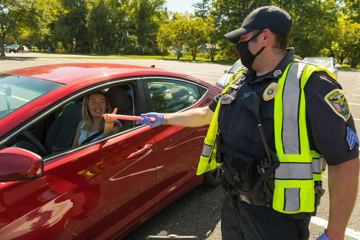 A police officer hands a safety item to a smiling woman in a red car at a campus parking lot.