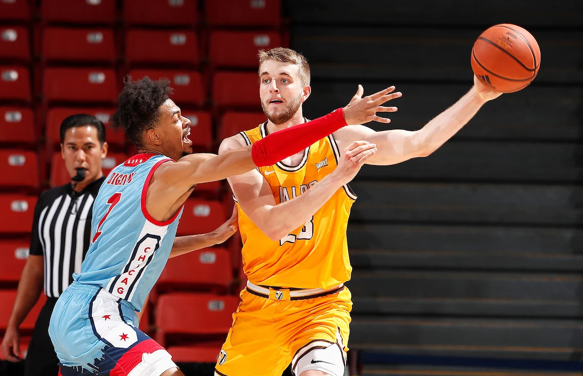Basketball player in yellow passes while being defended by a player in blue; the referee stands in the background during an exciting Uplift Valpo matchup.