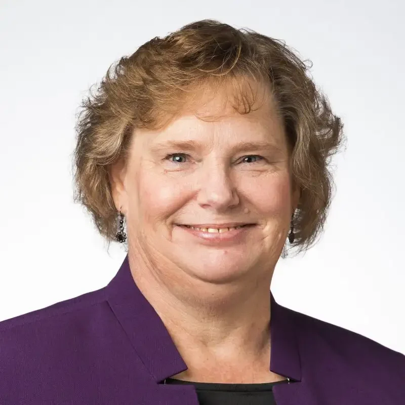 Smiling woman with short, curly hair wearing a purple blazer, photographed against a plain light background, exuding confidence and professionalism in communications.