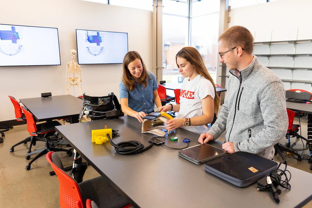 Three people explore electronics and gadgets on a table in a bright classroom with screens and red chairs, participating in a Peer Tutoring Program.