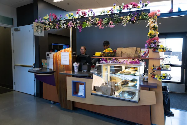 A small café counter decorated with colorful flowers, two staff working, and pastries displayed in a glass case creates a welcoming spot for health & wellness enthusiasts seeking fresh treats and vibrant atmosphere.