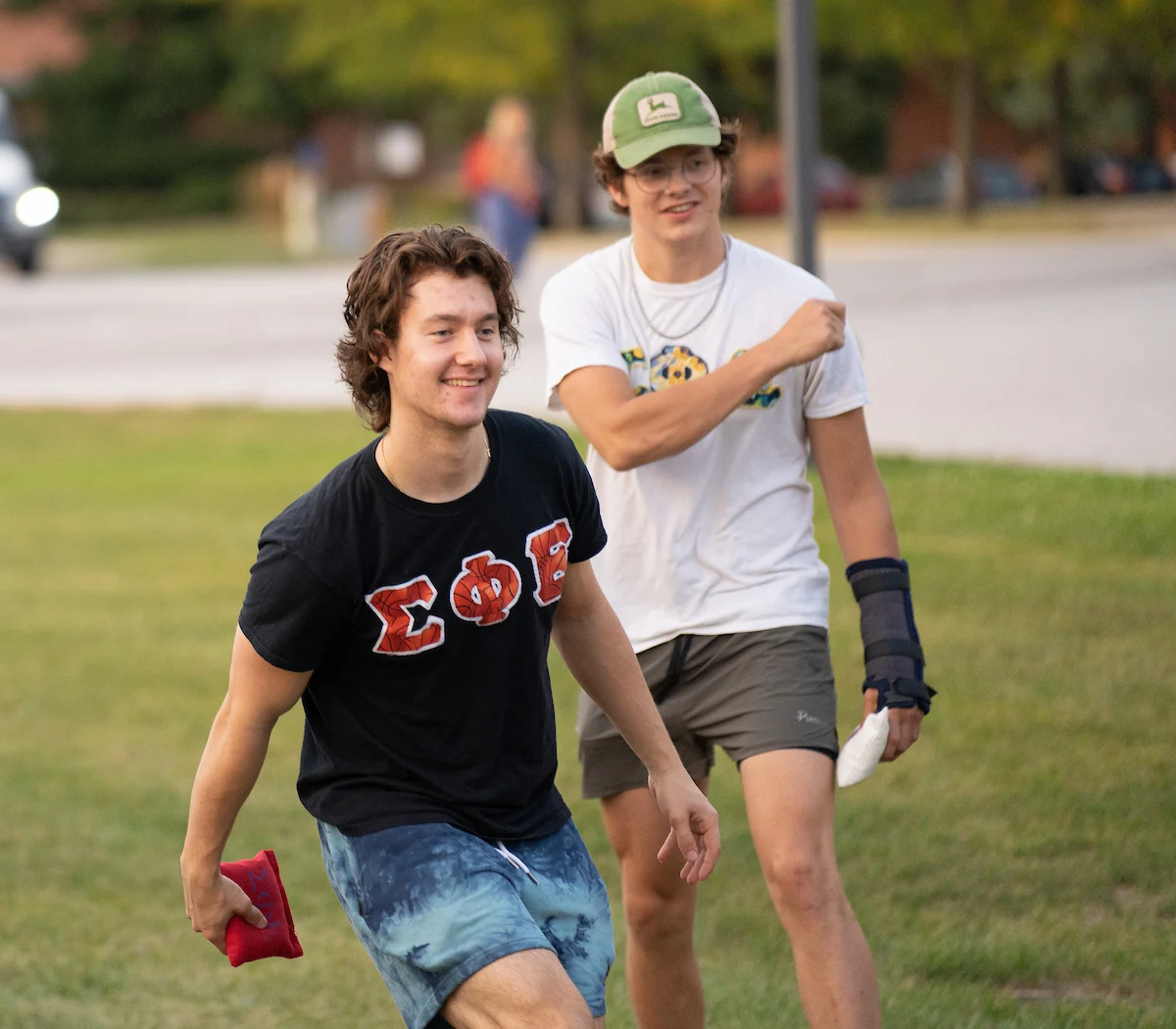 Two young men outside playing cornhole near a dining area; one smiles holding a bean bag, while the other has a cast on his right arm.