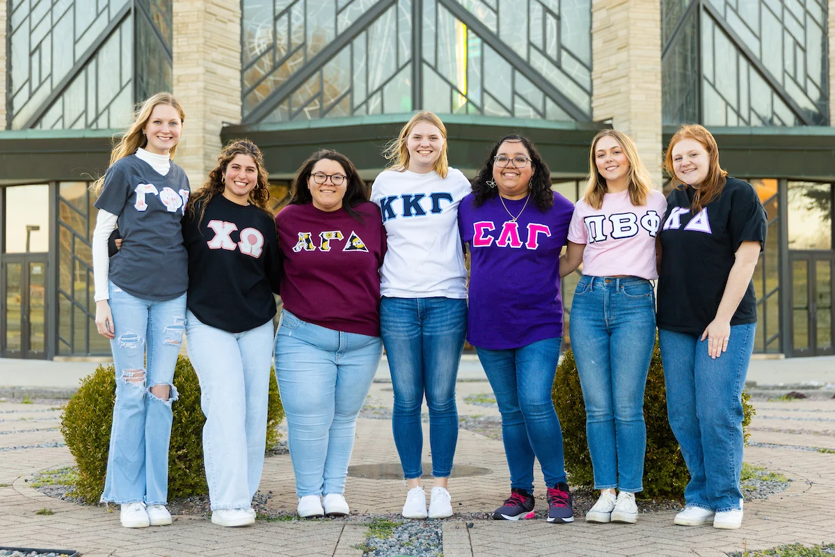 Seven smiling women in Greek letter shirts stand arm-in-arm outside a modern building, excited for a dining event together.
