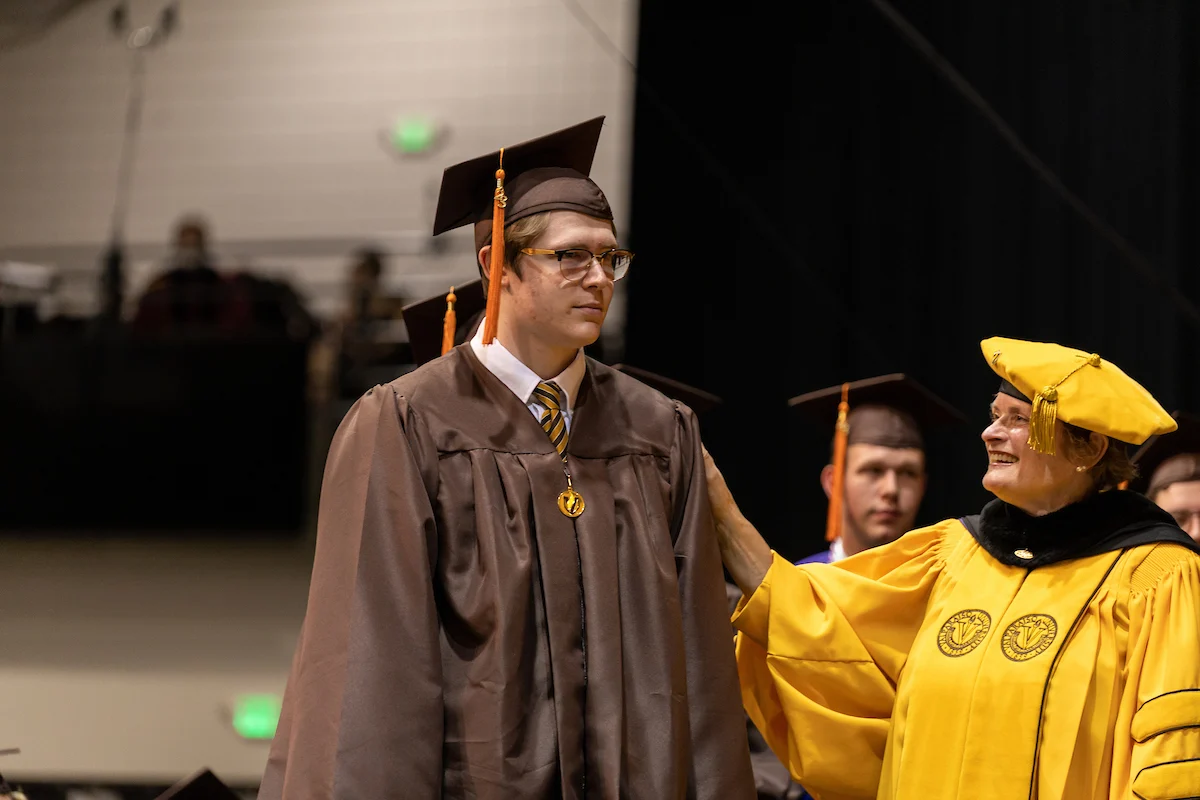 A graduate in a brown cap and gown stands next to a faculty member in a yellow robe at a ceremony, capturing an important moment highlighted in the Information for Graduates.