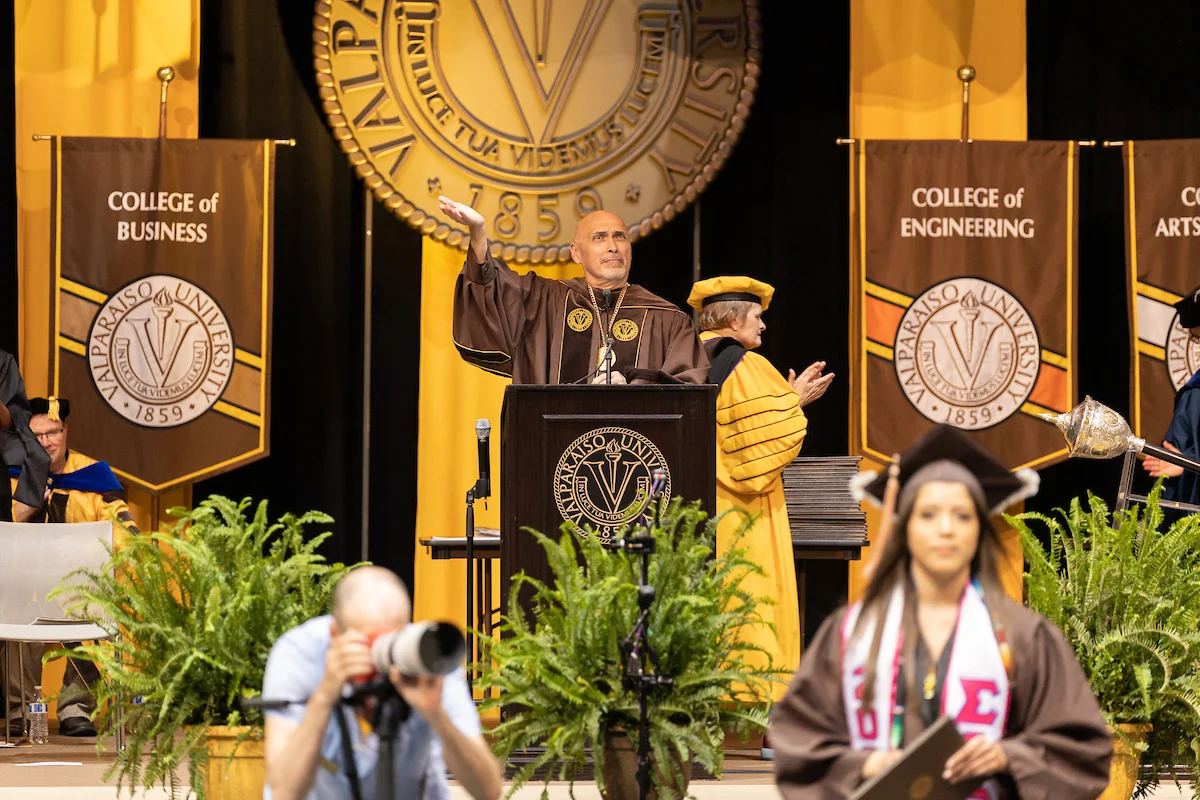 A man in academic regalia speaks at a graduation ceremony, sharing important Information for Graduates; graduates and photographers are in the foreground.