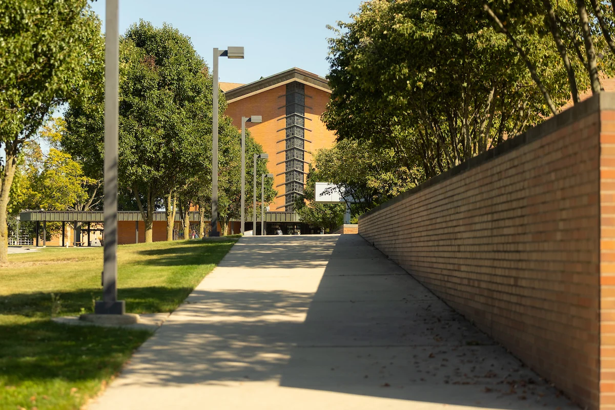 A concrete path lined with trees and a brick wall leads toward a modern brick building on a sunny day—¡Porque Somos Como Tú!.