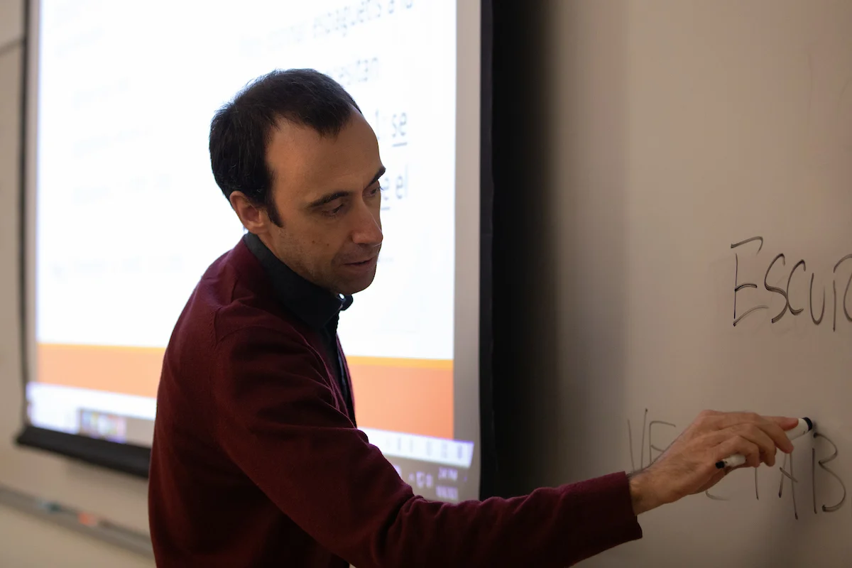 Man writing on a whiteboard in a classroom, with a projected presentation in the background highlighting Online Application Submission Forms.