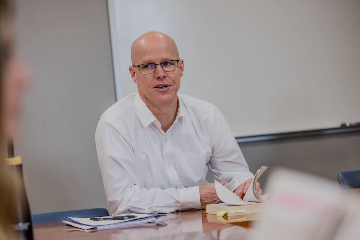 Bald man in glasses and white shirt sits at a table with papers, discussing Online Application Submission Forms in a meeting room.