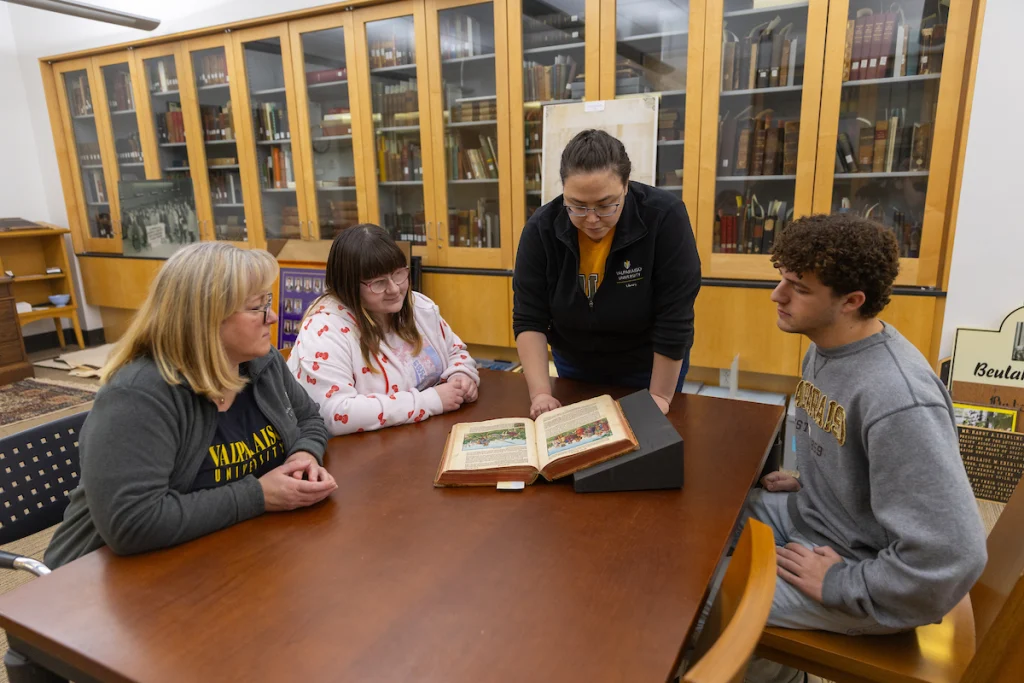 Four people sit at a table in a library or archive, examining an old book together as they discuss University Policies.