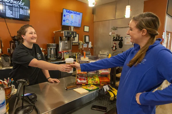 A barista hands a coffee cup to a smiling customer at a cafe counter featuring snacks and bananas, creating a warm atmosphere focused on Health & Wellness.