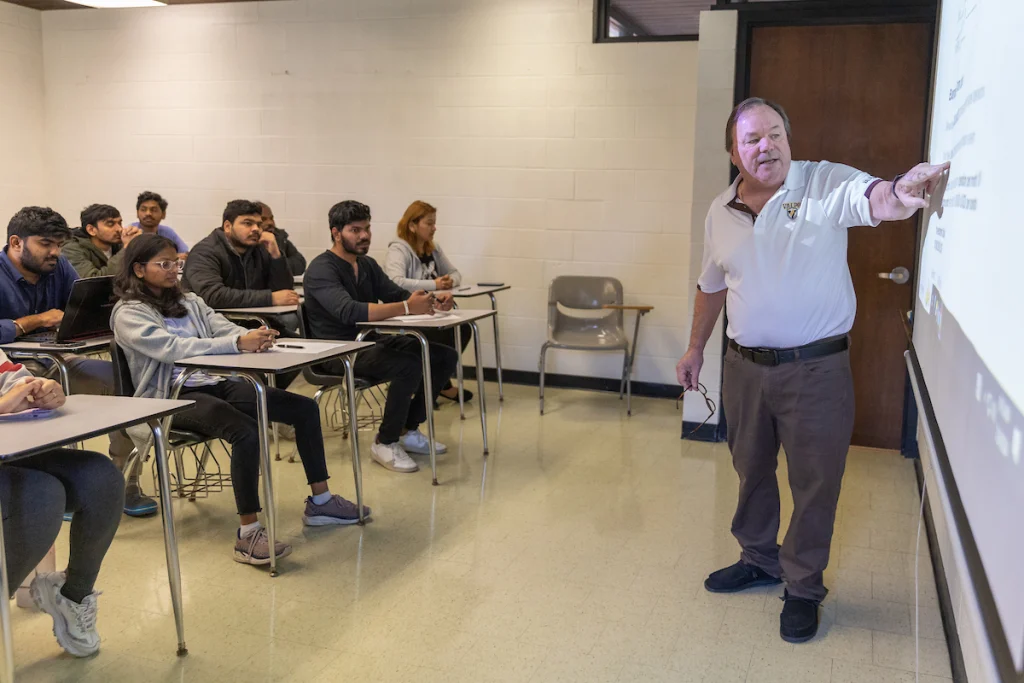 A teacher points at a screen while graduate students sit at desks and listen in a classroom.