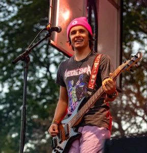 Smiling musician Zach Stewart in a pink beanie and Metallica shirt playing guitar on an outdoor stage at Valpo, radiating a sense of belonging.