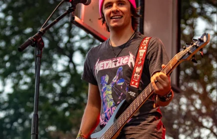 Smiling musician Zach Stewart in a pink beanie and Metallica shirt playing guitar on an outdoor stage at Valpo, radiating a sense of belonging.