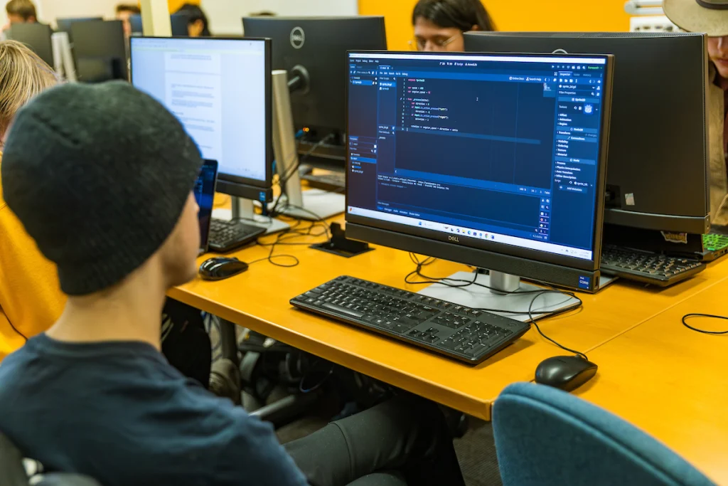 Person wearing a beanie working on code at a desktop computer in a classroom, contributing to the collaborative spirit of Uplift Valpo with others engaged in the background.