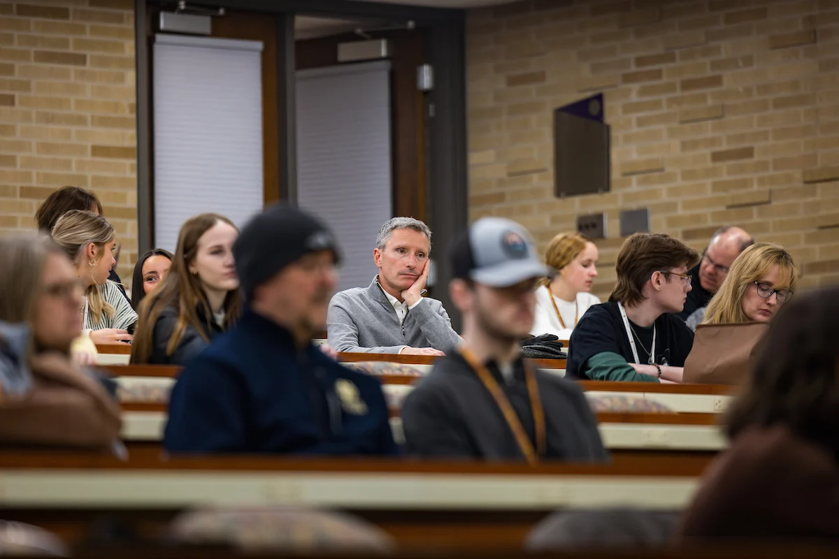 People sit and listen with focus in a university lecture hall, surrounded by rows of seats and brick walls.