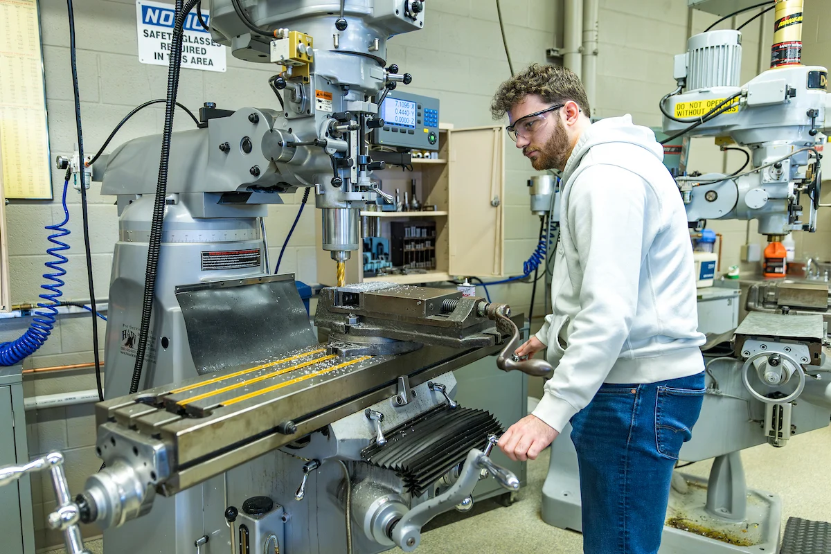 A man in safety glasses operates a milling machine in a workshop, demonstrating hands-on experience that admitted students can look forward to.