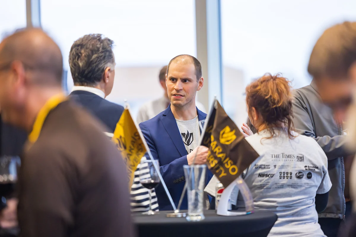 A man in a blue blazer talks with others at a social event, holding a Valparaiso University flag to Uplift Valpo spirit.
