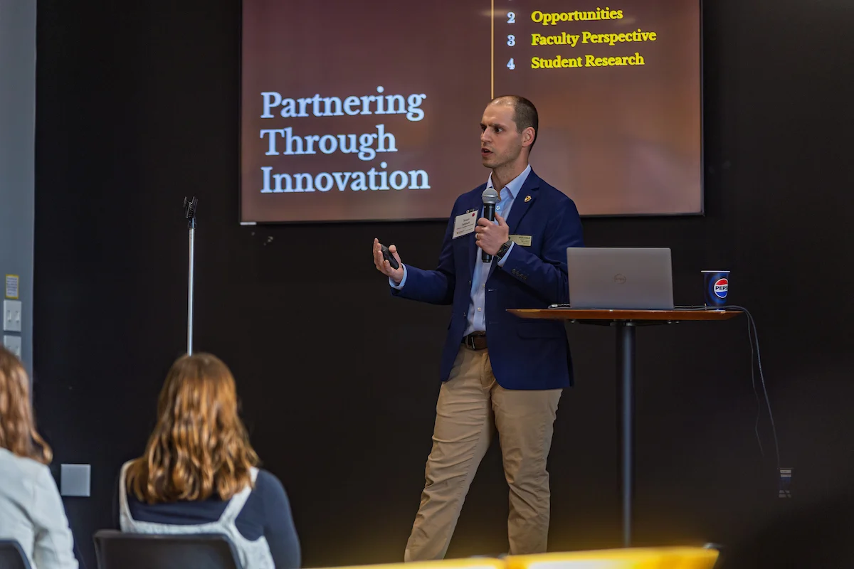 A man in a suit speaks at a podium with a PowerPoint slide reading "Partnering Through Innovation" during a Committee to Enhance Learning & Teaching event.