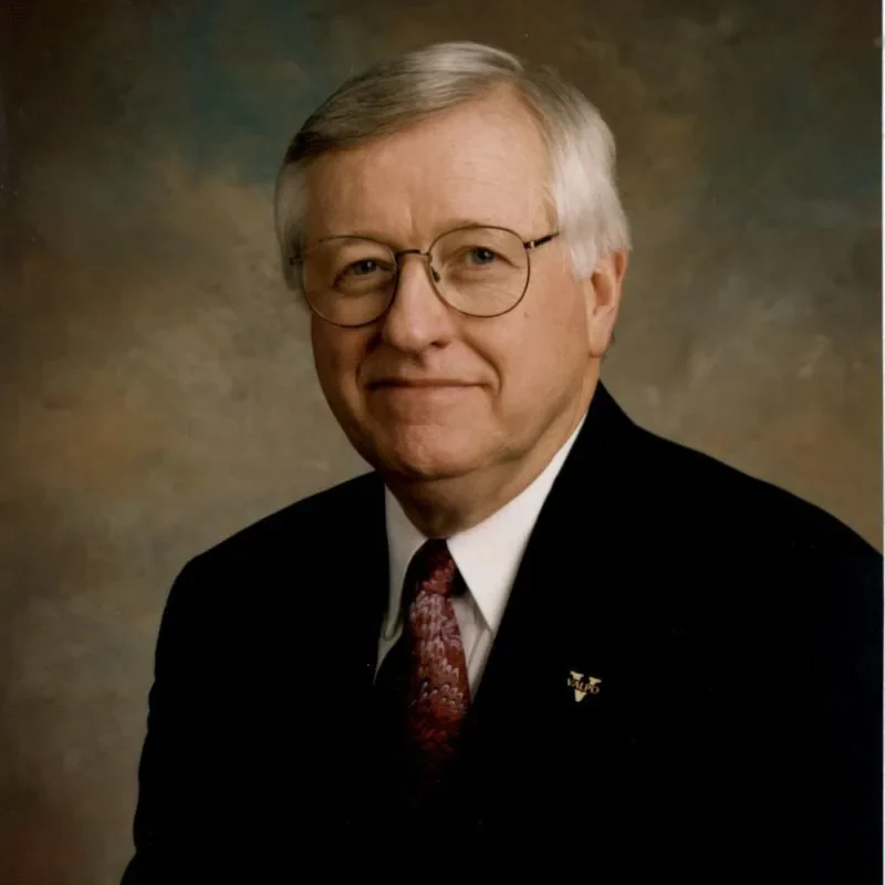 Older man with gray hair and glasses wearing a suit, tie, and pin, embodying leadership and governance as he poses against a neutral background.