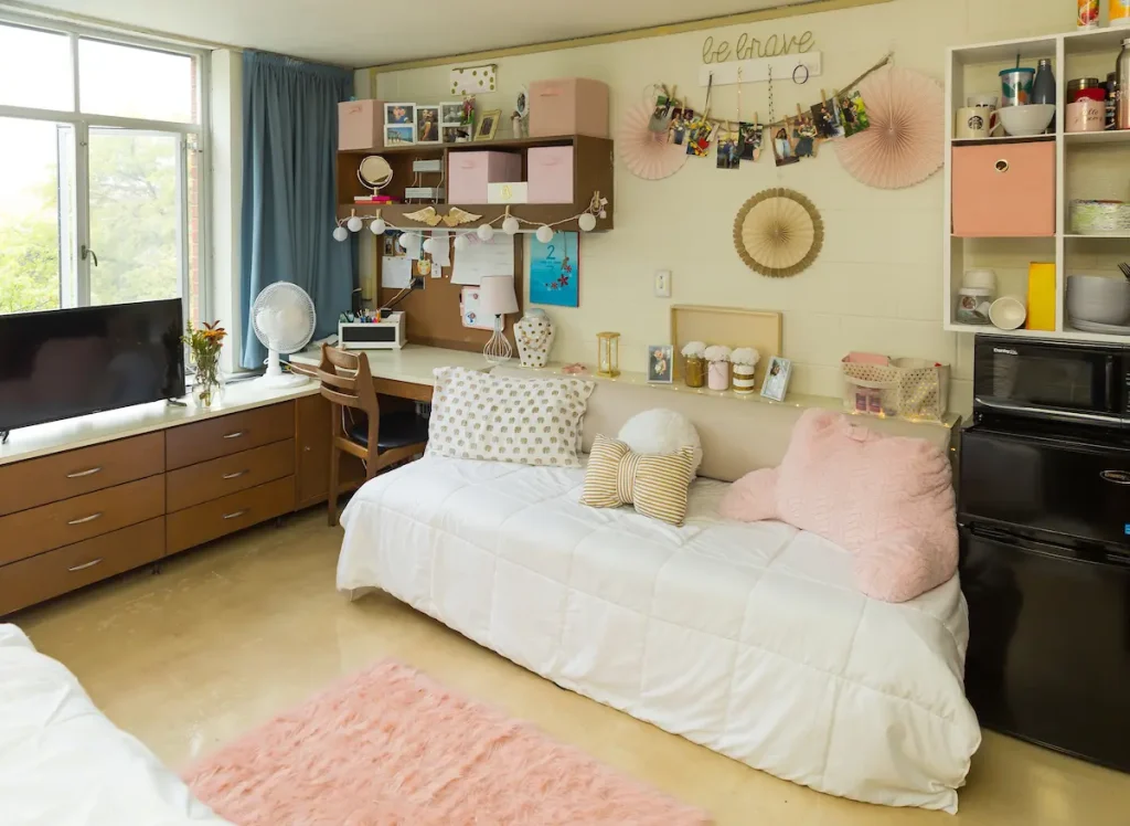 A cozy dorm room with a bed, pillows, desk, shelves, photos, and a TV. Decor is light and pastel-colored.