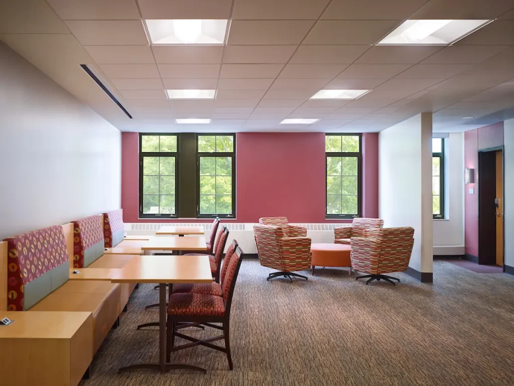 Modern lounge with tables, cushioned chairs, large windows, and natural light in a quiet, empty room.