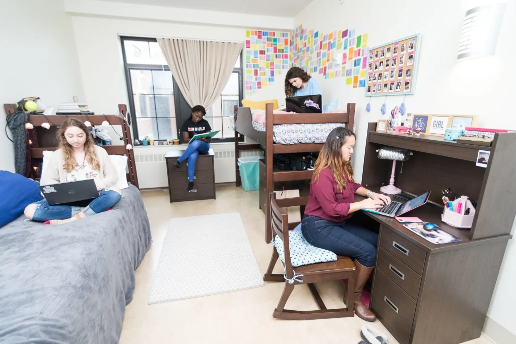 Four young women studying on laptops in a bright, colorful college dorm room with lofted beds and desks.