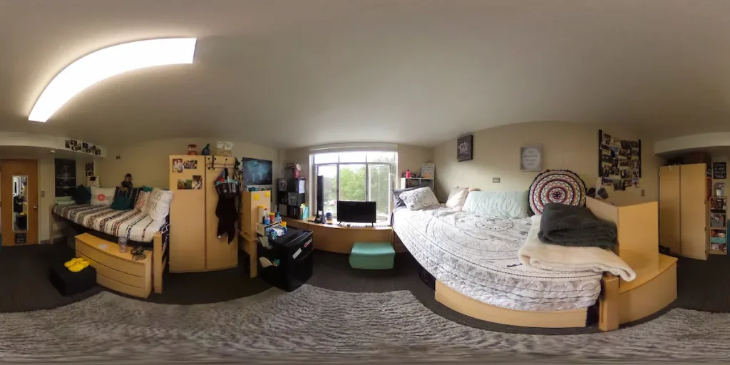 A wide-angle view of a dorm room with two beds, desks, shelves, and a window in the center.