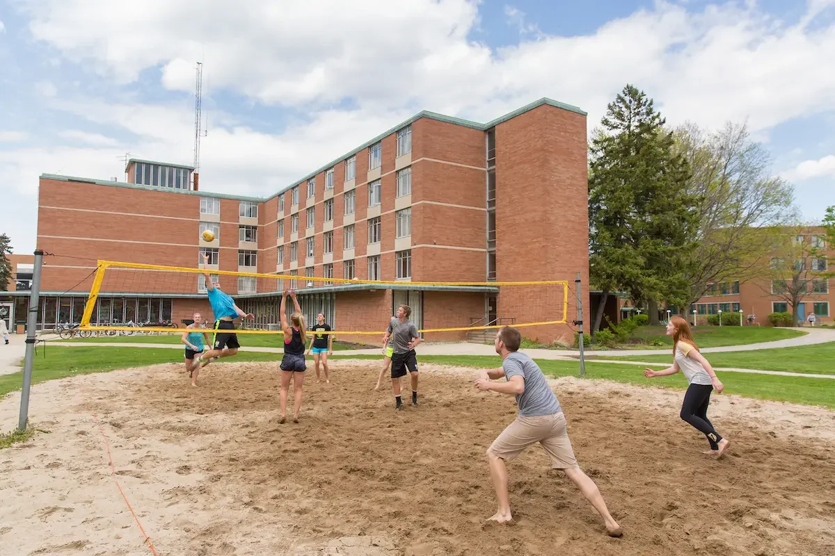 Six people play beach volleyball on a sandy court outside a brick dormitory building on a sunny day.