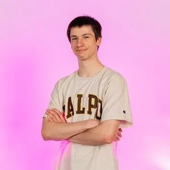 Young person with short dark hair, arms crossed, wearing a "VALPO" shirt, standing in front of a pink background—representing the dedicated Student Support Assistants (SSAs) team.