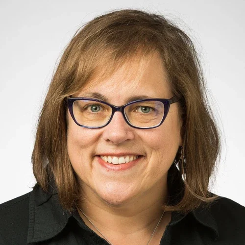 Smiling woman with shoulder-length light brown hair and glasses, wearing a black shirt, on a plain background, reflecting the warmth and openness of Our Lutheran Identity.