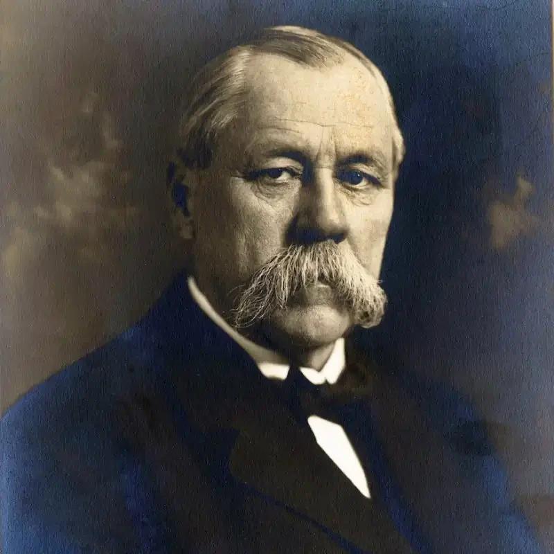 An older man with a large mustache, embodying leadership and governance, wears a suit and bow tie as he poses for a formal portrait.