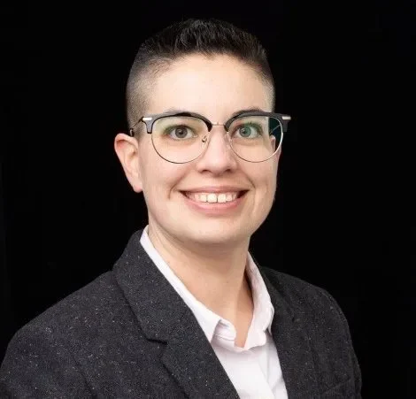 Person with short dark hair, glasses, and a blazer smiling against a black background, radiating confidence in the field of communications.
