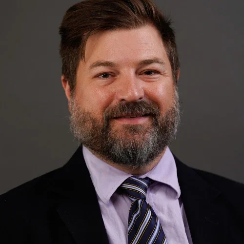 Man with brown hair and beard wearing a suit, light purple shirt, and striped tie, smiling, reflecting the warmth and professionalism of Our Lutheran Identity.