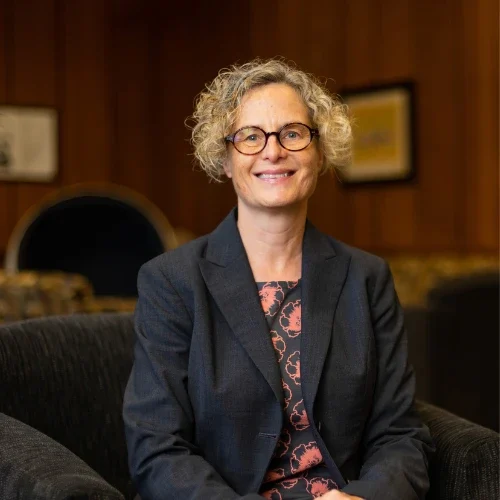 Smiling person with curly hair and glasses sits in a chair, wearing a suit in a wood-paneled room that reflects our Lutheran Identity.