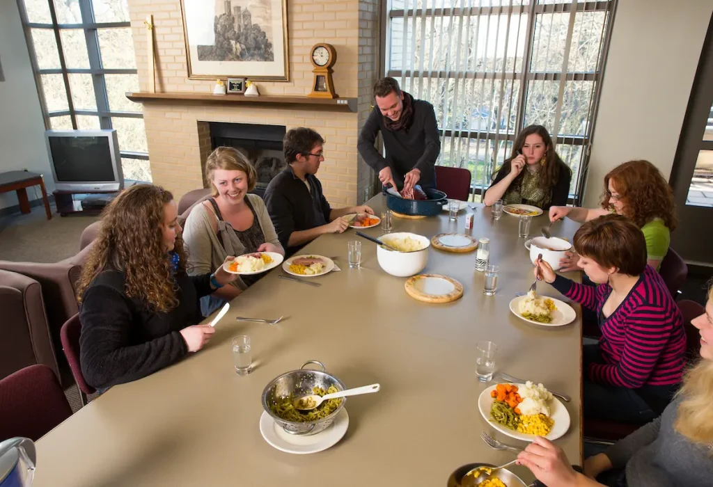 Eight people sit around a table enjoying a meal together in the cozy, sunlit dining room of the German House, reflecting the warmth and community of German language housing.
