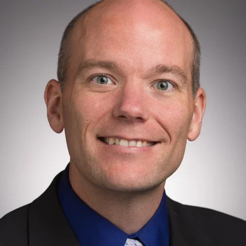 Smiling man in a suit and blue shirt, with short hair, against a plain gray background, reflecting confidence and embodying our Lutheran identity.