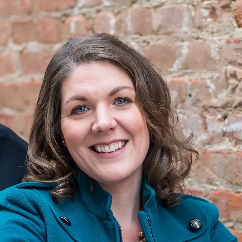 Smiling woman with wavy brown hair, wearing a teal coat, stands in front of a brick wall—radiating confidence and communications expertise.